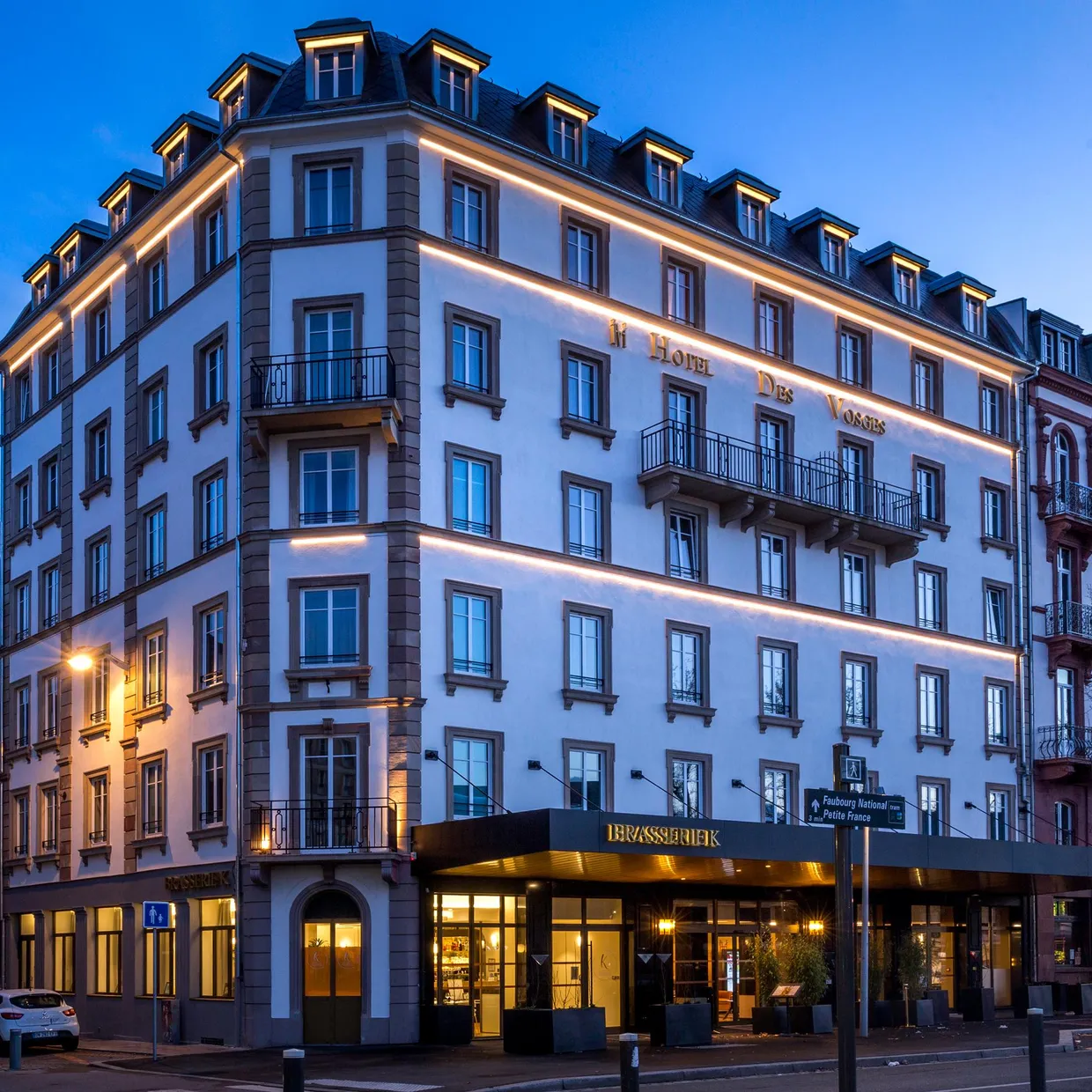 Eckgebäude nachts beleuchtet mit der Aufschrift "Hotel Des Voyageurs" und einem Restaurant im Erdgeschoss
