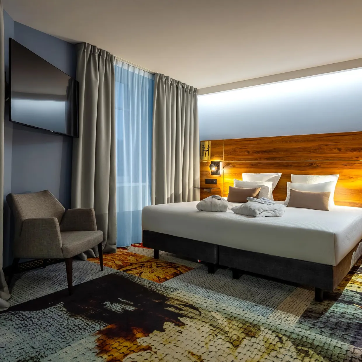 Hotelzimmer mit einem Doppelbett, zwei Sesseln, einem Holzkopfteil und grauen Vorhängen