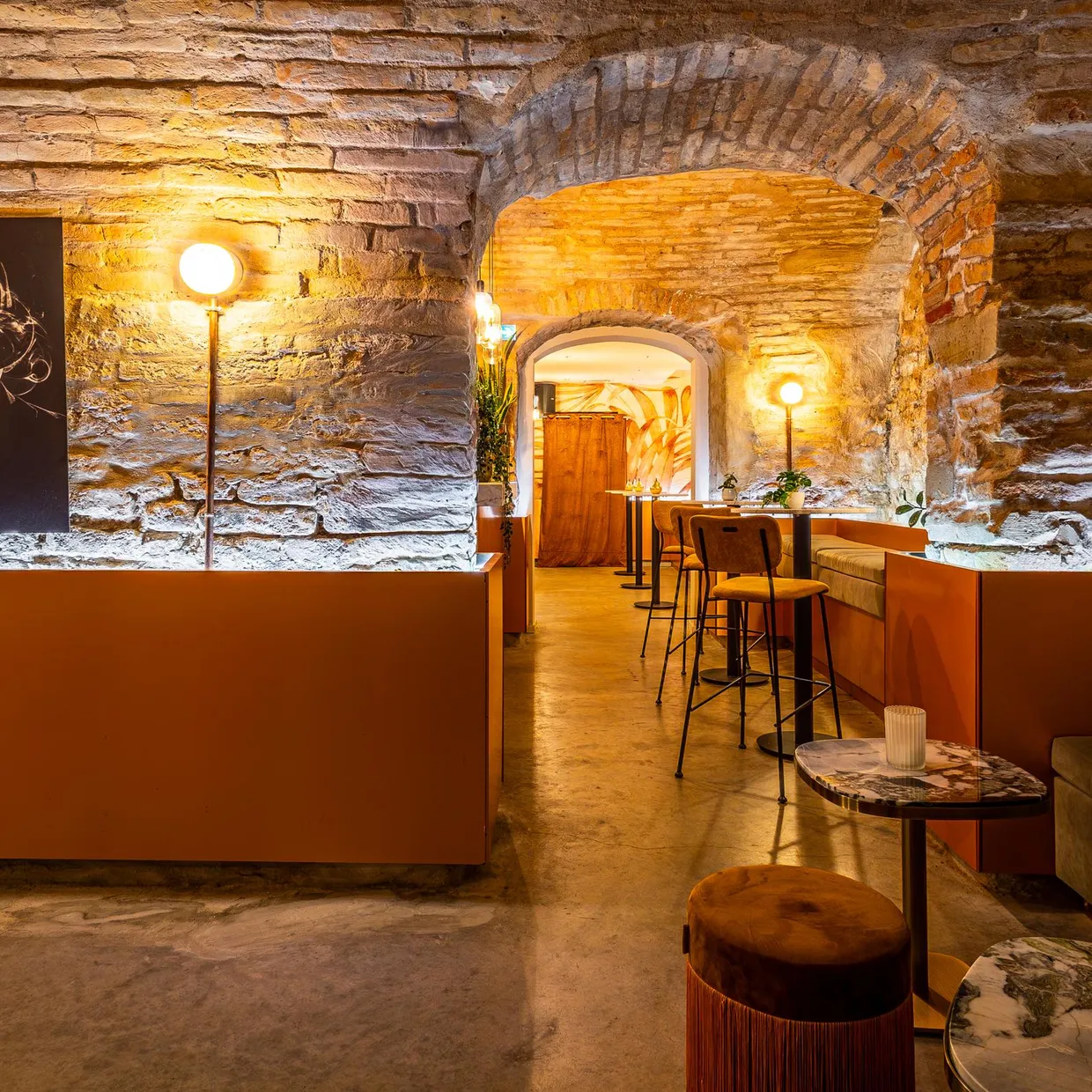 Ein Innenraum eines Bars oder Restaurants mit freiliegenden Steinwänden, Stehlampen, Tischen und Bänken, beleuchtet durch warmes Licht