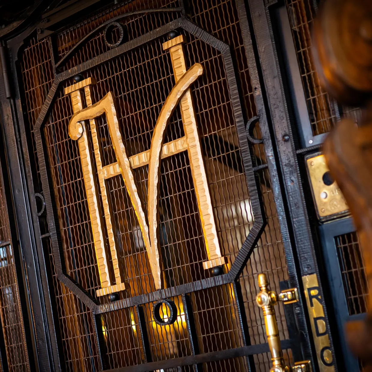 Metallgitter mit den erhabenen Buchstaben "M" und "A", beleuchtet von goldenen Lichtern, in einem dunklen architektonischen Rahmen