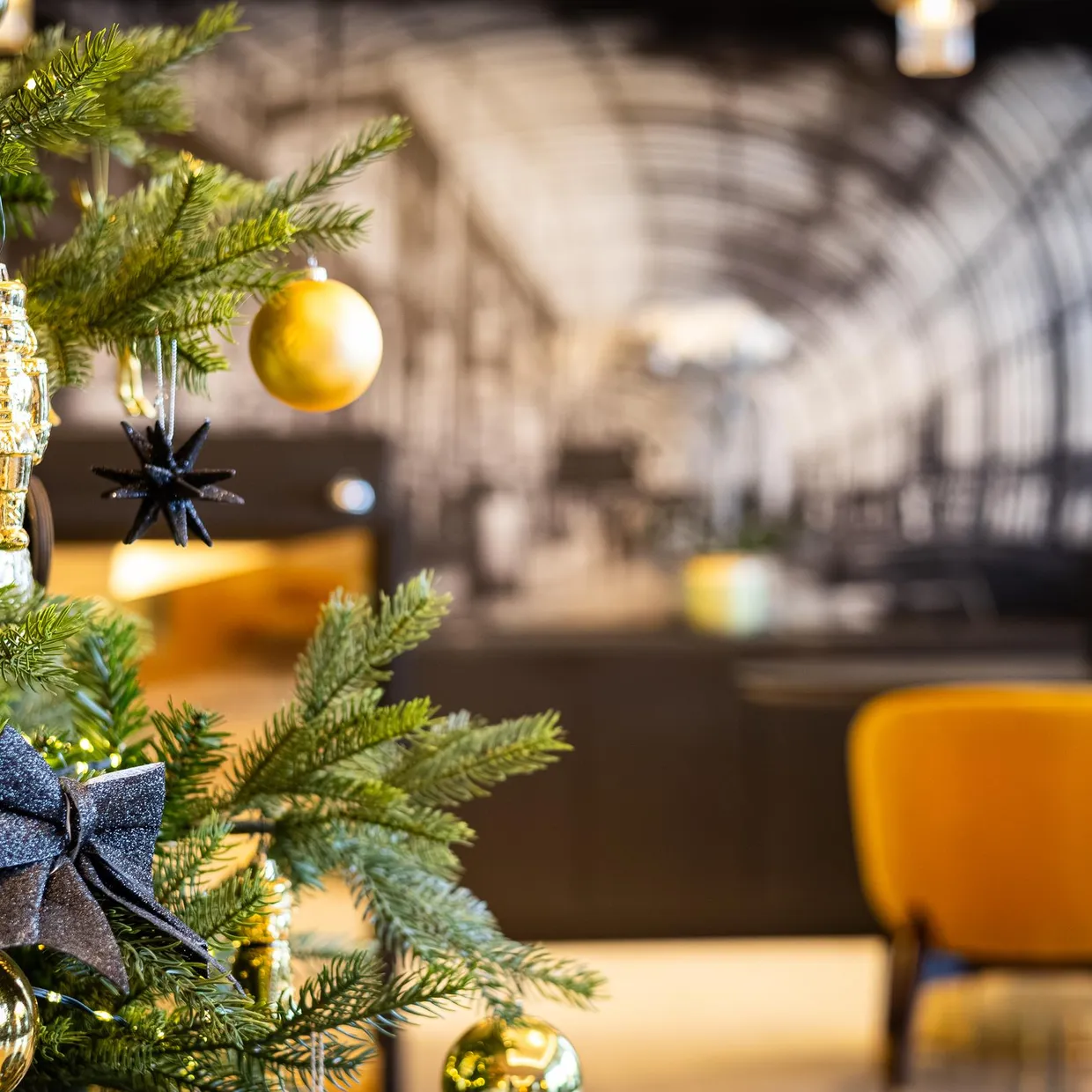 Ein mit goldenen und schwarzen Verzierungen geschmückter Weihnachtsbaum in einem Innenraum mit gelben Stühlen und einer Ziegelwand