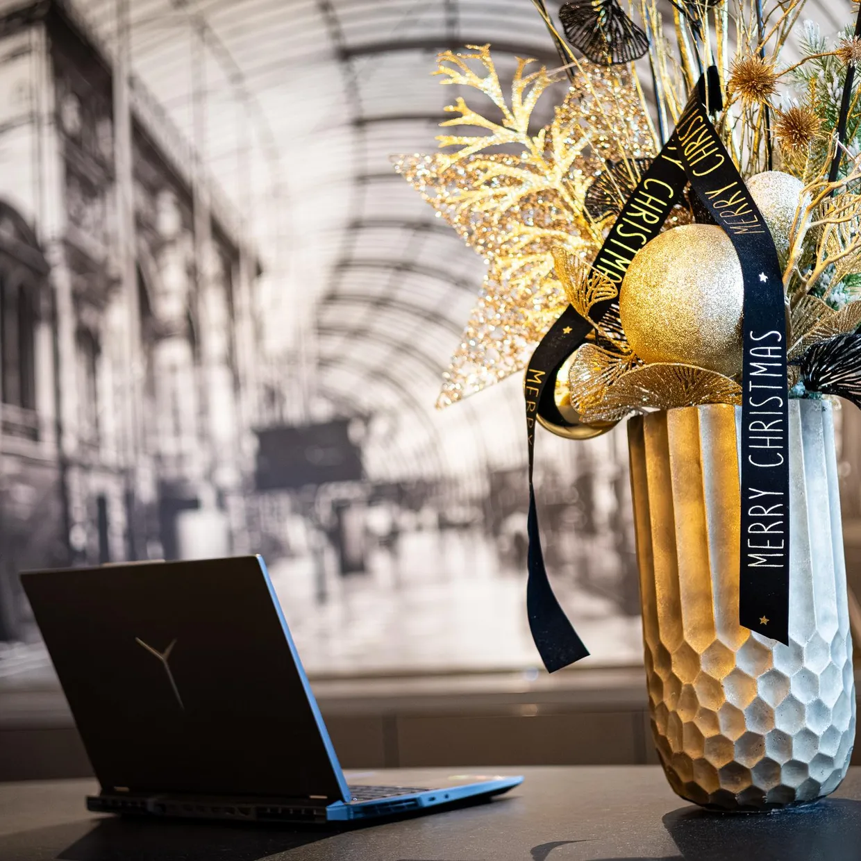 Eine mit festlichen Elementen verzierte Vase mit einem "Merry Christmas"-Band steht auf einem Tisch neben einem Laptop vor einer großen Glasfront.