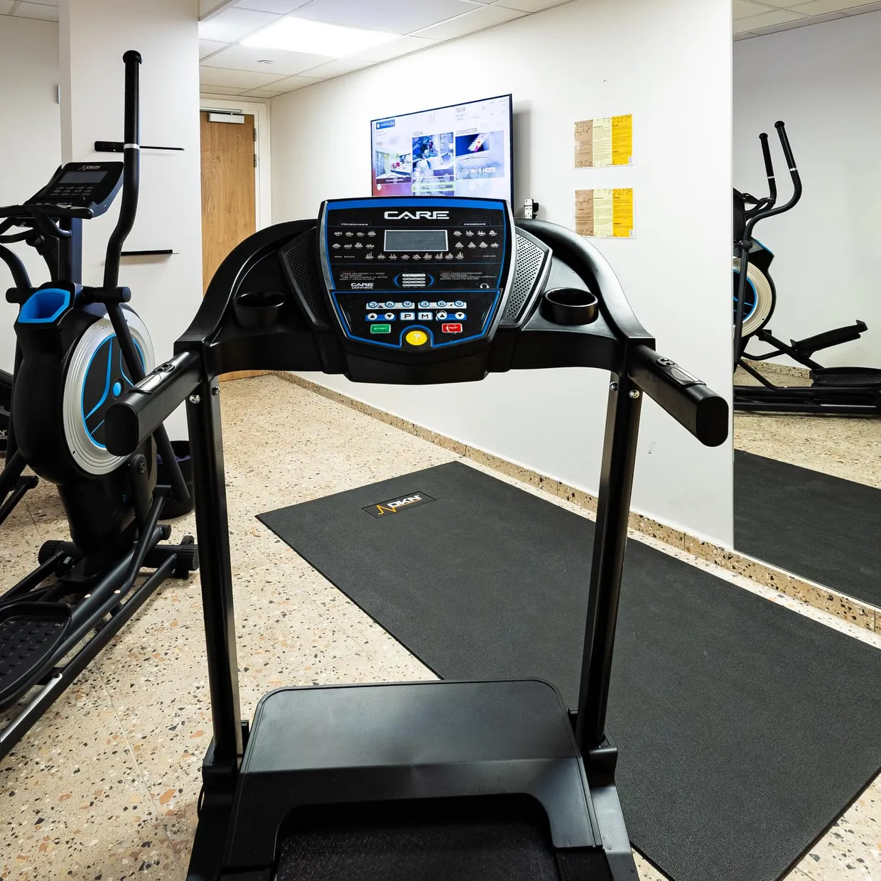 Fitnessraum mit mehreren Cardio-Geräten, darunter ein Laufband und zwei Ellipsentrainer, auf Bodenmatten platziert