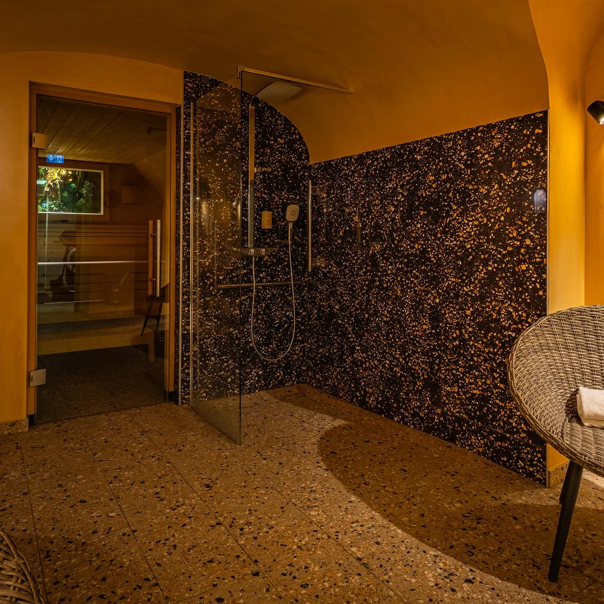 Duschraum mit schwarzer und goldener Mosaikwand, zwei Rattanstühlen und einer offenen Tür zu einem angrenzenden Raum