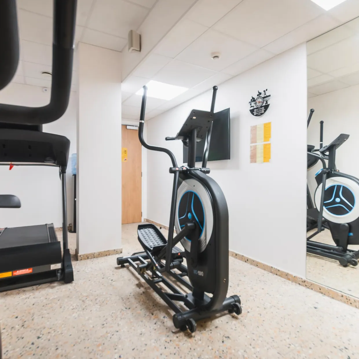 Fitnessraum mit mehreren Cardio-Geräten, darunter ein Laufband und Ellipsentrainer, vor einem Spiegel