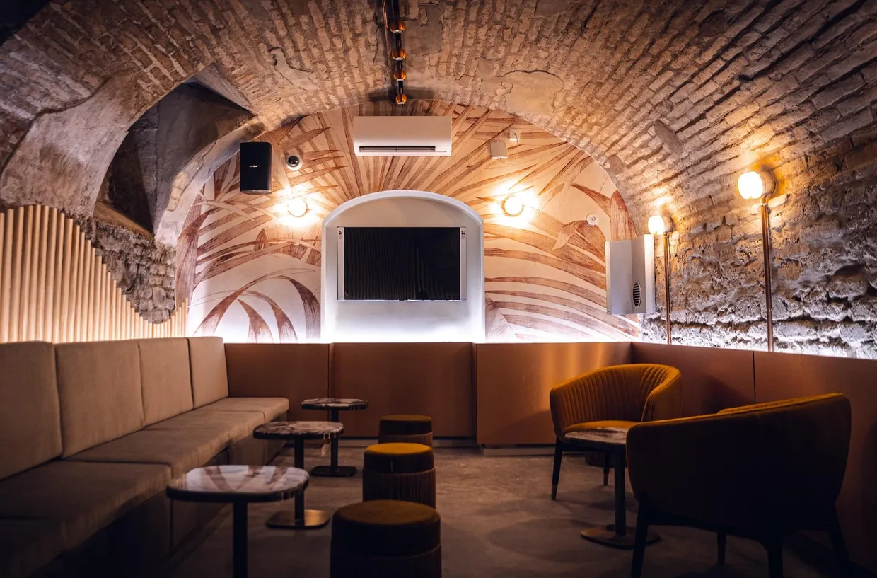Underground room with stone arches, beige sofa, yellow armchairs, and screen at the back
