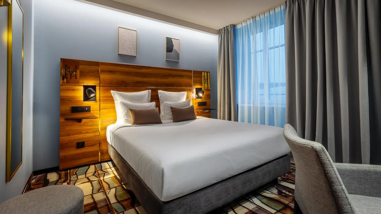 Hotelzimmer mit einem großen Doppelbett, weißen Kissen und braunen Polstern, einem Holzkopfteil mit integrierter Beleuchtung, blauen Vorhängen und einem Fenster, das Tageslicht hereinlässt.