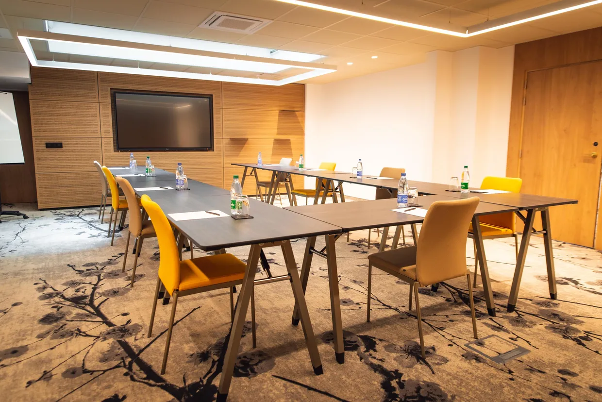 Meeting room with tables and chairs arranged in a U-shape, equipped with a screen and water bottles on each table.