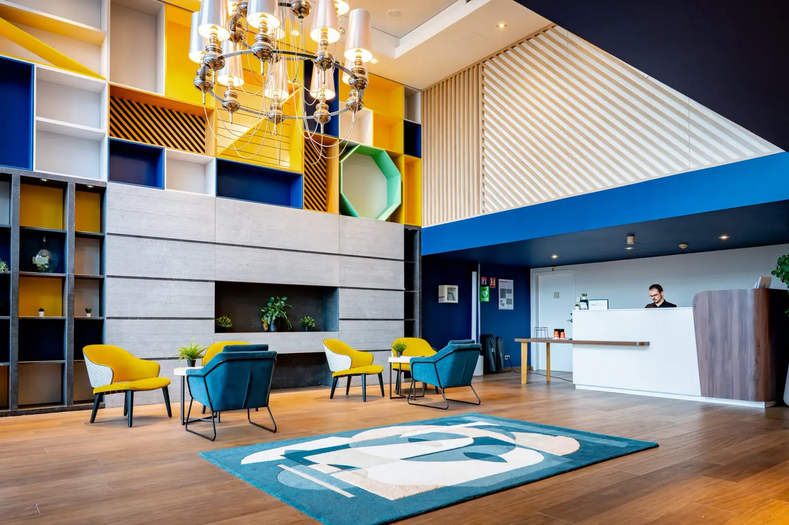 Moderne Hotelrezeption mit bunten Stühlen, einem Couchtisch, einem Empfangstresen und einem hängenden Kronleuchter