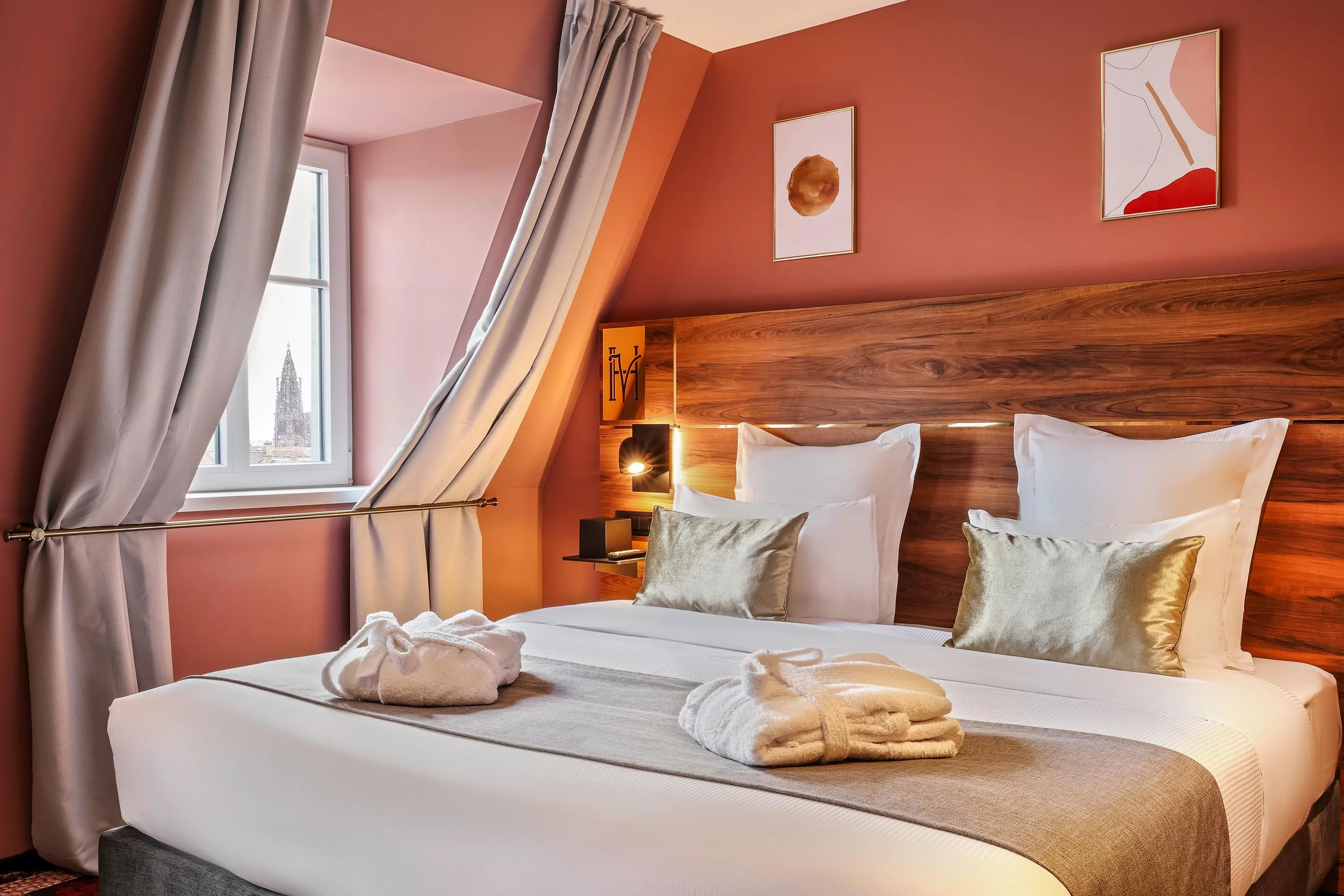 Zimmer mit einem Doppelbett, Kissen und Decken, einem Holzkopfteil, grauen Vorhängen und einem Fenster mit Blick auf einen Turm.