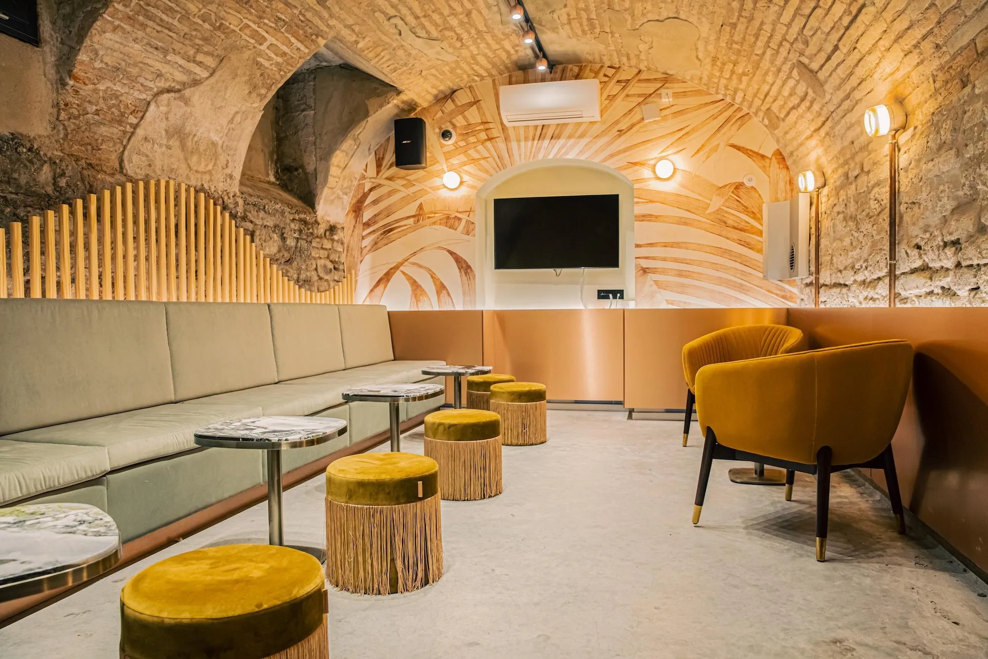 Ein Innenraum mit Steinbögen, einer mit Holzmustern verzierten Decke, einem beigen Sofa, Hockern und Sesseln mit gelbem Samt sowie einem an der Wand montierten Fernseher