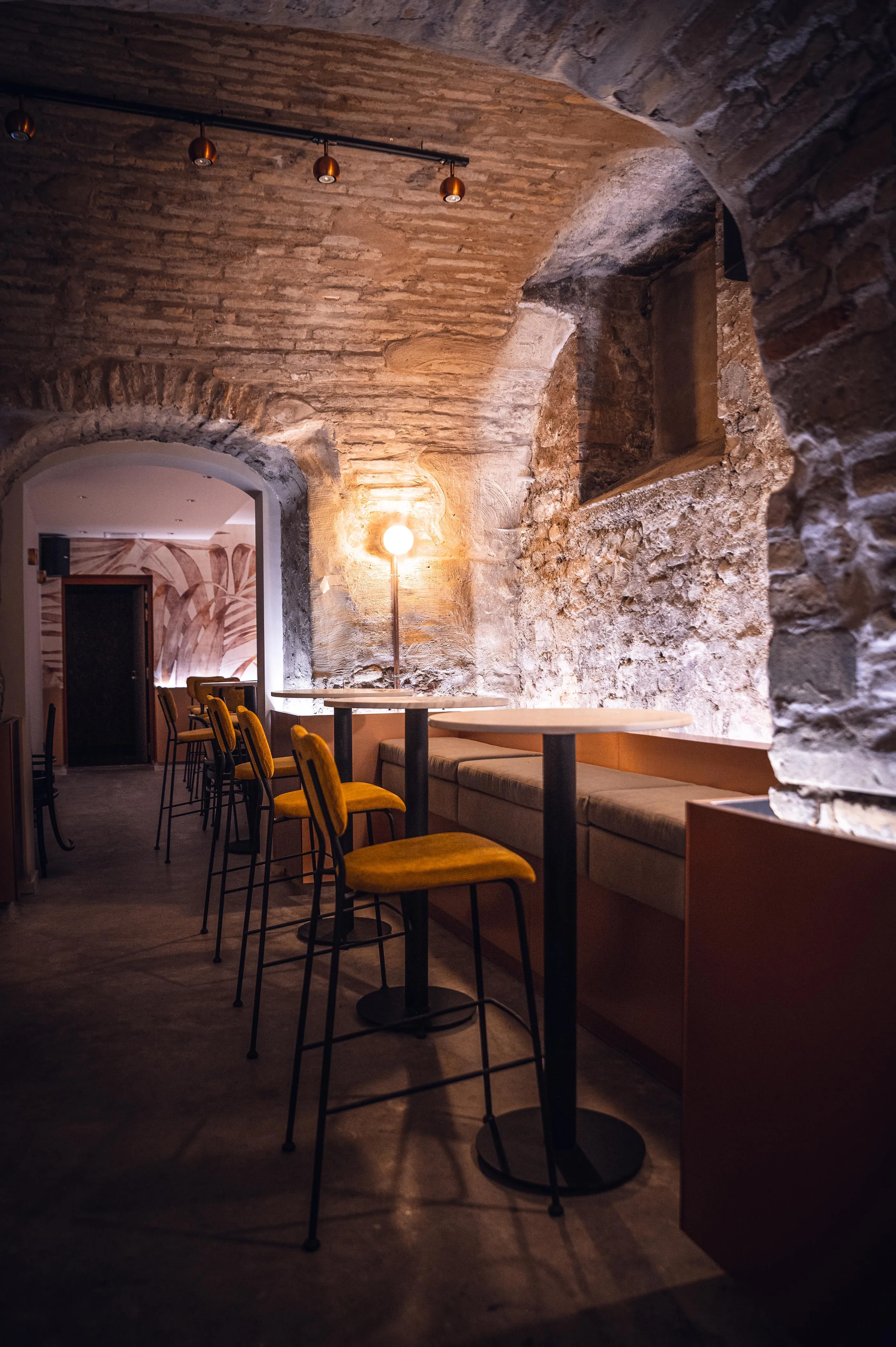 Eine Bar mit gelben Hockern und einem hohen Tisch, die sich in einem Raum mit Steinwänden befindet, der von Lampen beleuchtet wird