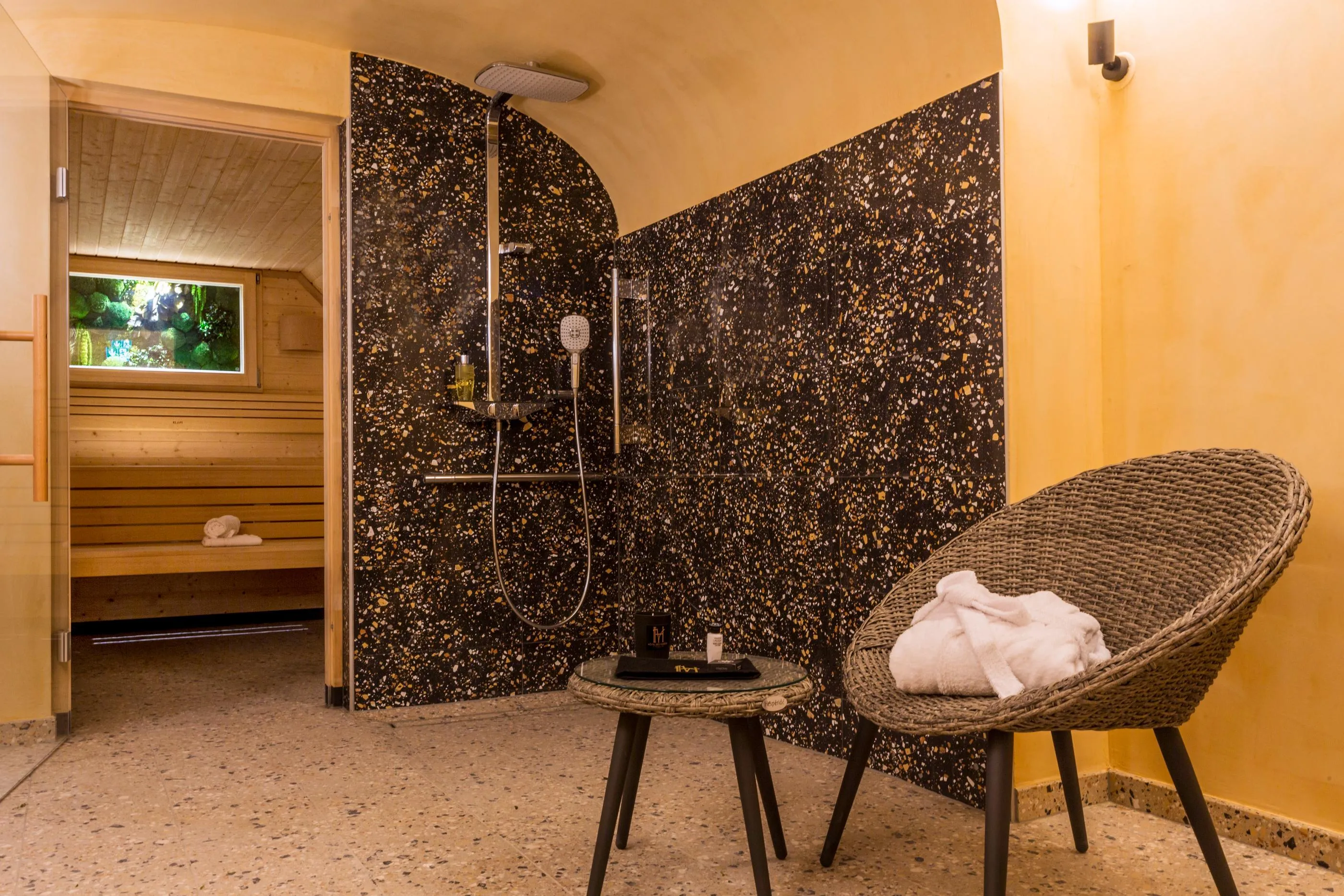 Badezimmer mit Wanddusche, schwarzer und goldener Kabine, Korbstuhl mit weißem Handtuch und Couchtisch