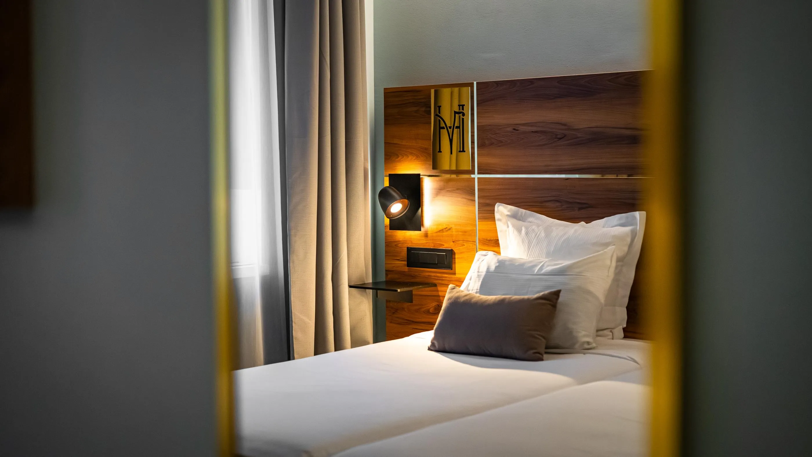 Bett mit Holz-Kopfteil, weißen Kissen und einem grauen Kissen, beleuchtet von einer Nachttischlampe