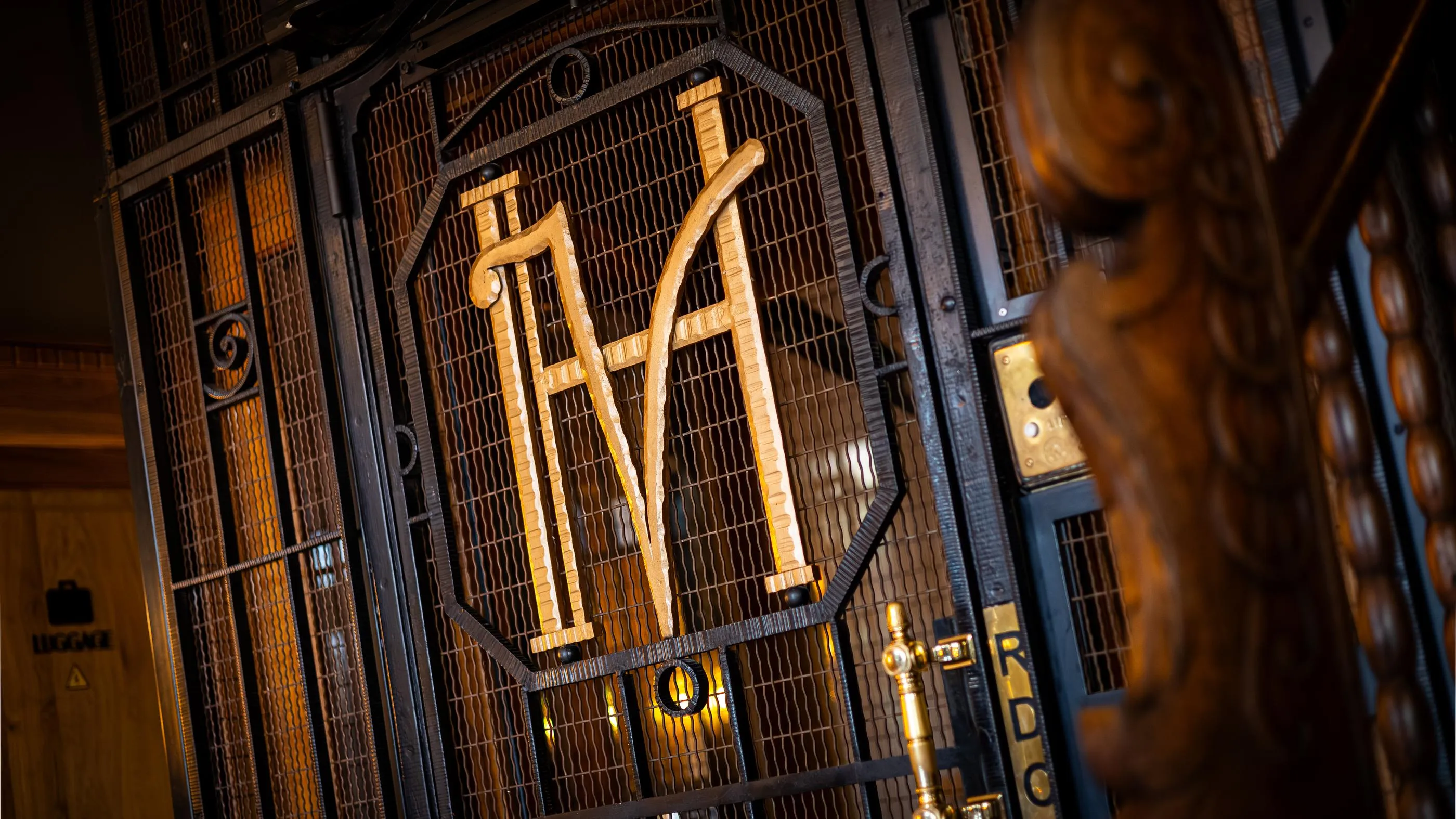 Metallgitter mit den erhabenen Buchstaben "M" und "A", beleuchtet von goldenen Lichtern, in einem dunklen architektonischen Rahmen