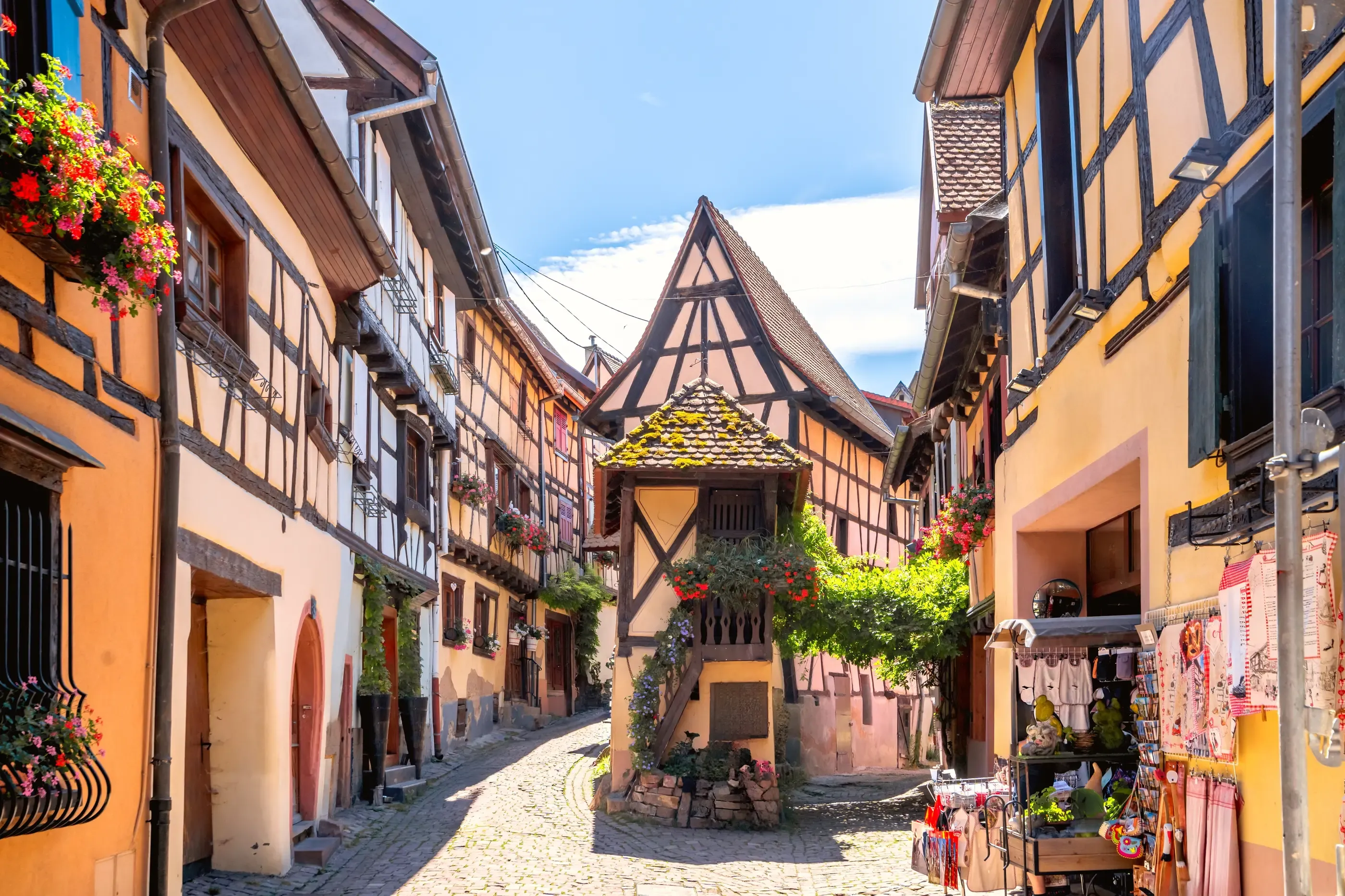 Mit Kopfsteinpflaster gepflasterte Straße, gesäumt von farbenfrohen Fachwerkhäusern mit hängenden Blumen und einem kleinen Holzkiosk in der Mitte.