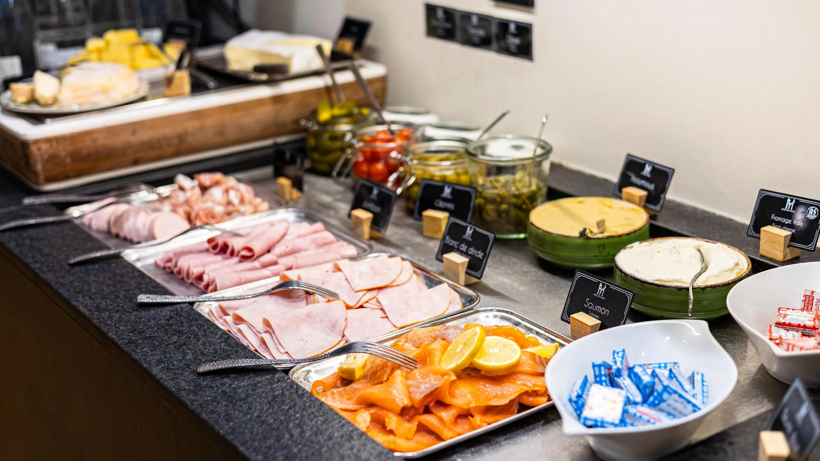 Ein Buffet mit verschiedenen Fleisch- und Wurstwaren, Käsen, eingelegten Gemüsesorten und Räucherlachs, auf einem Tisch mit Holzetiketten angerichtet