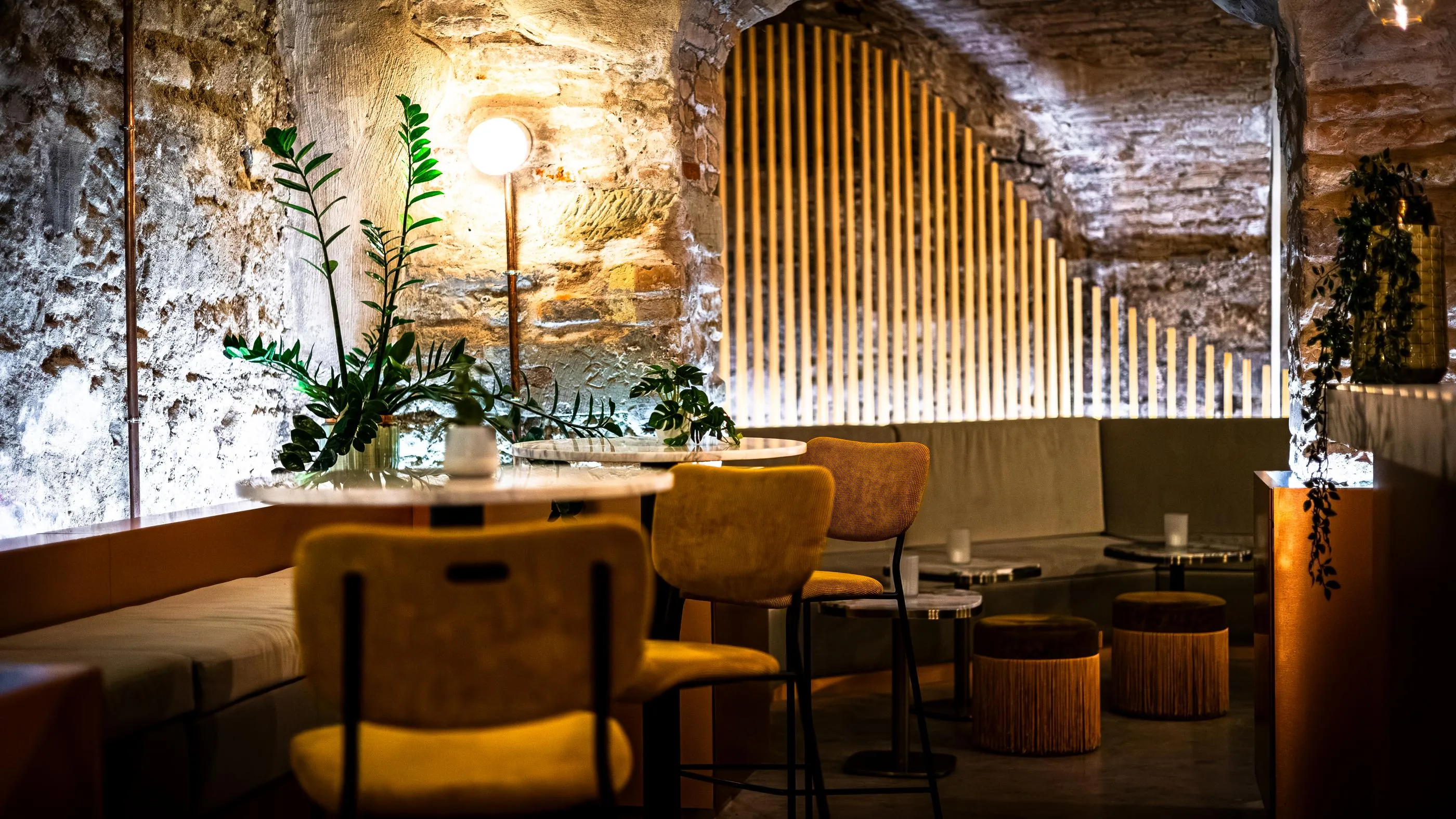 A café interior with stone walls, tables, chairs, and benches, lit by candles and green plants.
