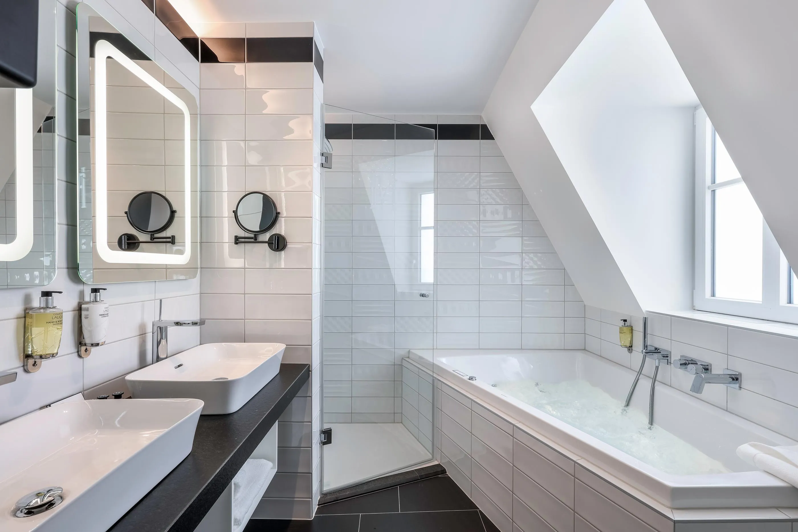 Modernes Badezimmer mit zwei Waschbecken, einer mit Wasser gefüllten Badewanne und einer Dusche, alles in Weiß und Schwarz gefliest