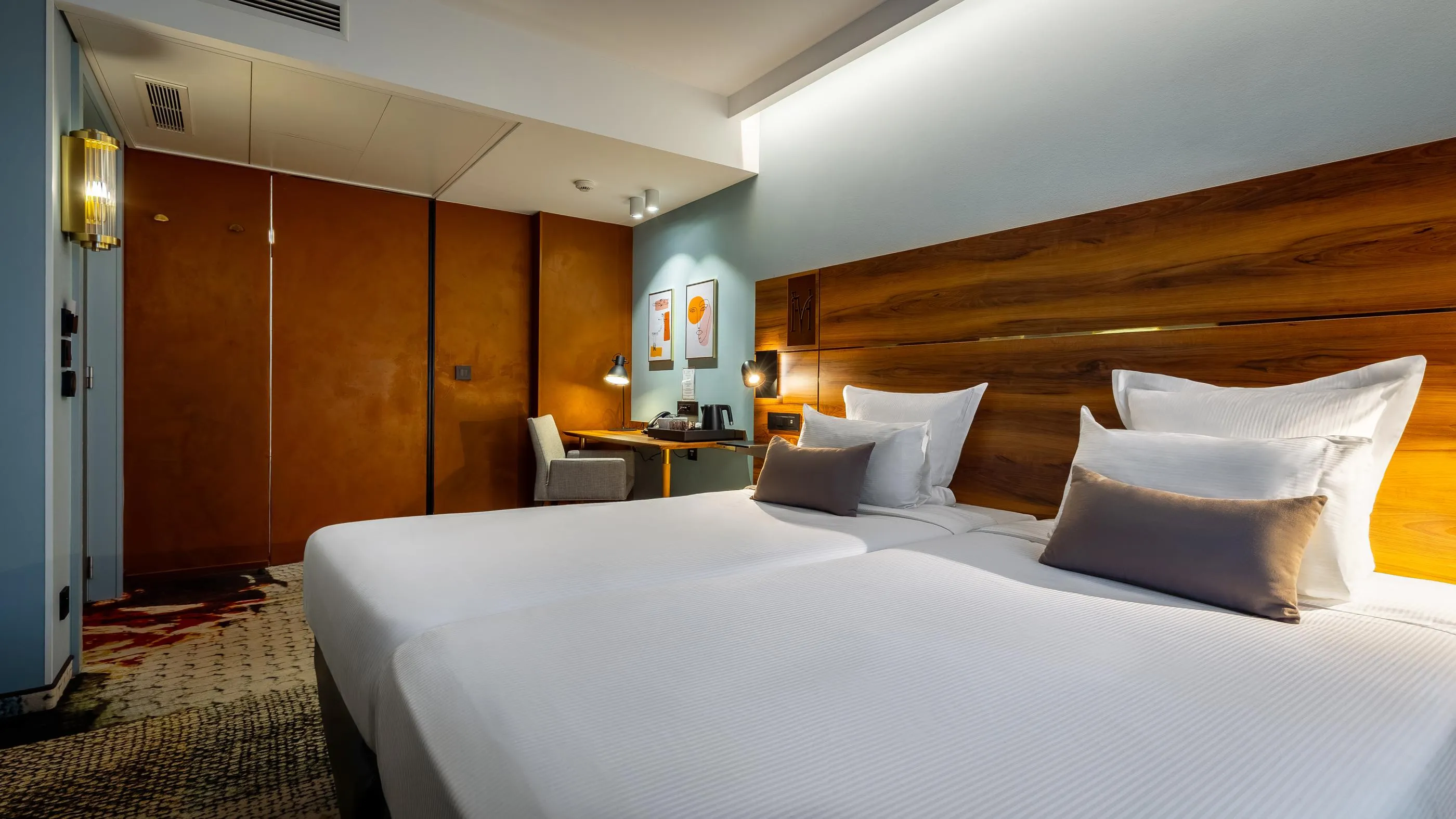 Hotel room with two single beds, a wooden headboard, a desk with a chair, and artwork on the wall.