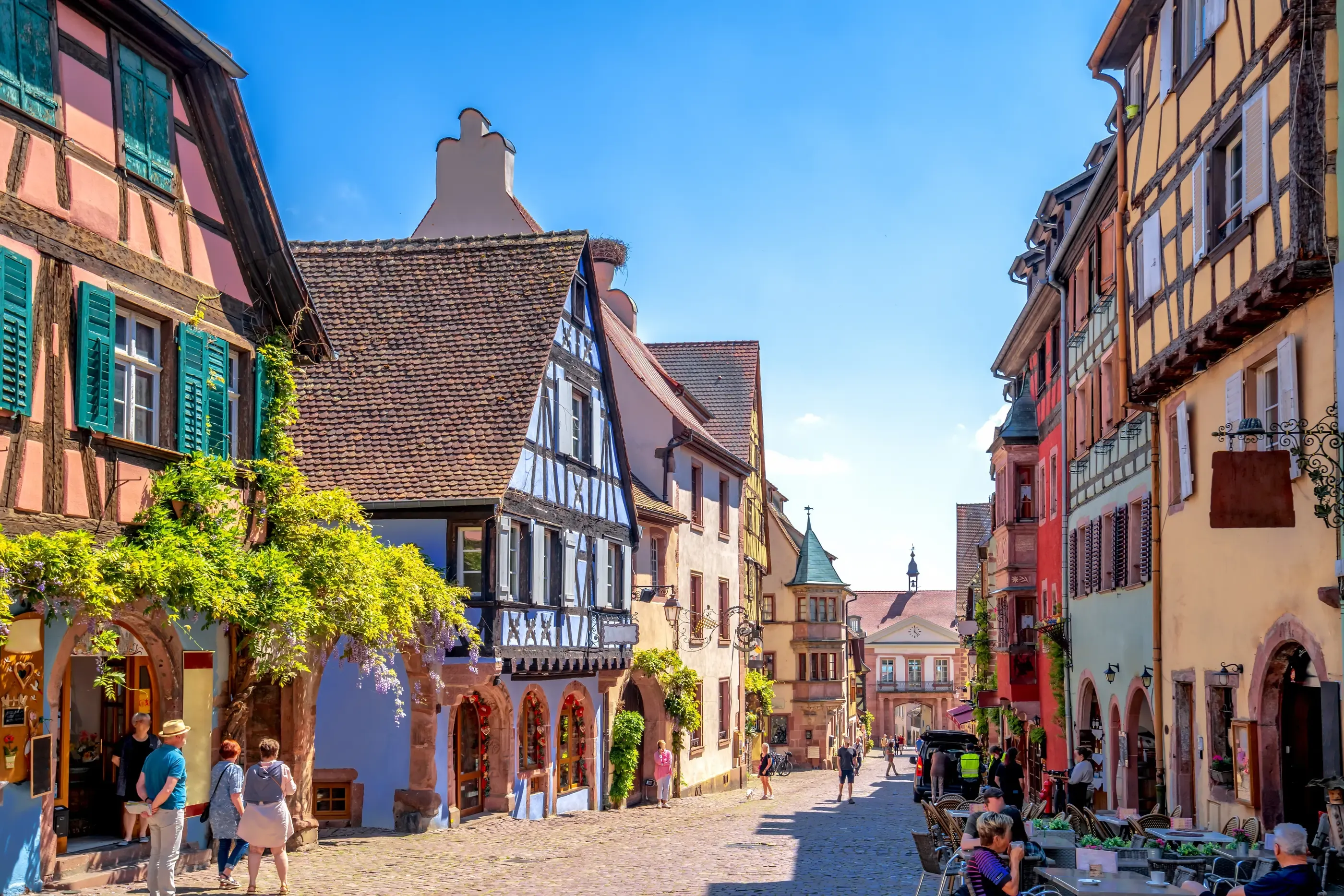 Mit Kopfsteinpflaster gepflasterte Straße, gesäumt von farbenfrohen Fachwerkhäusern, mit Menschen, die gehen, und Café-Tischen auf der Terrasse.