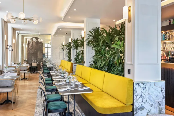 Elegant restaurant with set tables, green chairs, and a yellow banquette, green plants, and a visible marble bar at the back.