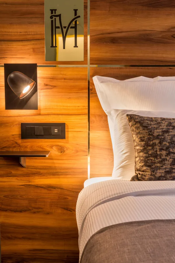Bett mit Holzkopfteil, eingeschalteter Wandlampe und weißen und grauen Kissen