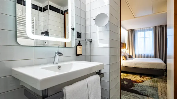 Modernes Badezimmer mit weißem Waschbecken, beleuchtetem Spiegel und offener Tür zum Schlafzimmer