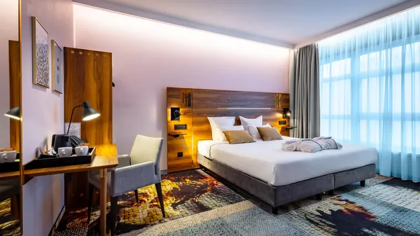 Hotelzimmer mit einem Doppelbett, einem Schreibtisch, einem Stuhl und großen Fenstern, die Tageslicht hereinlassen
