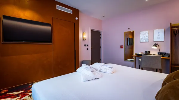 Hotelzimmer mit einem mit Handtüchern bedeckten Bett, einem Wandfernseher, einem Schreibtisch mit einer Lampe und Kunstwerken an der Wand