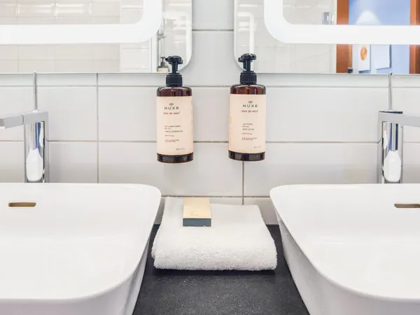 Two Nuxe "rêve de miel" product dispensers suspended between two sinks with towels and soap