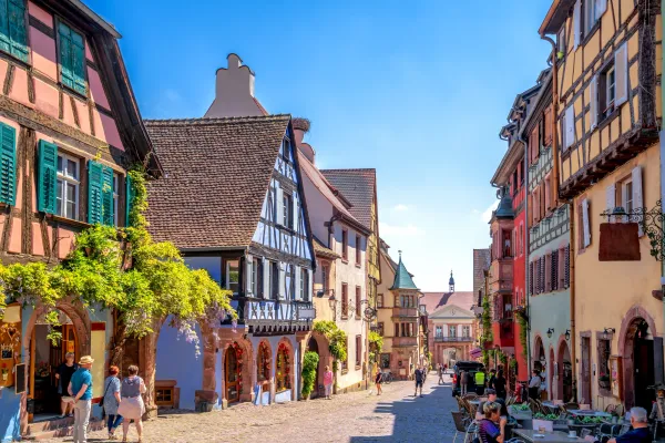 Cobbled street lined with colorful timber-framed houses, with people walking and café tables on the terrace.